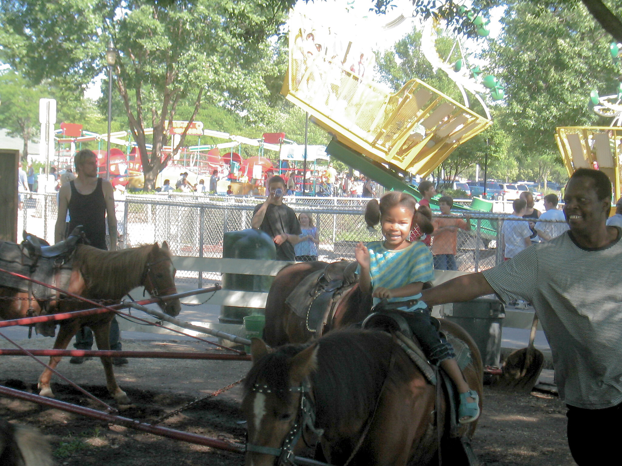 Children ride a yellow carnival carousel with other rides and trees in the background; a fence is in the foreground.