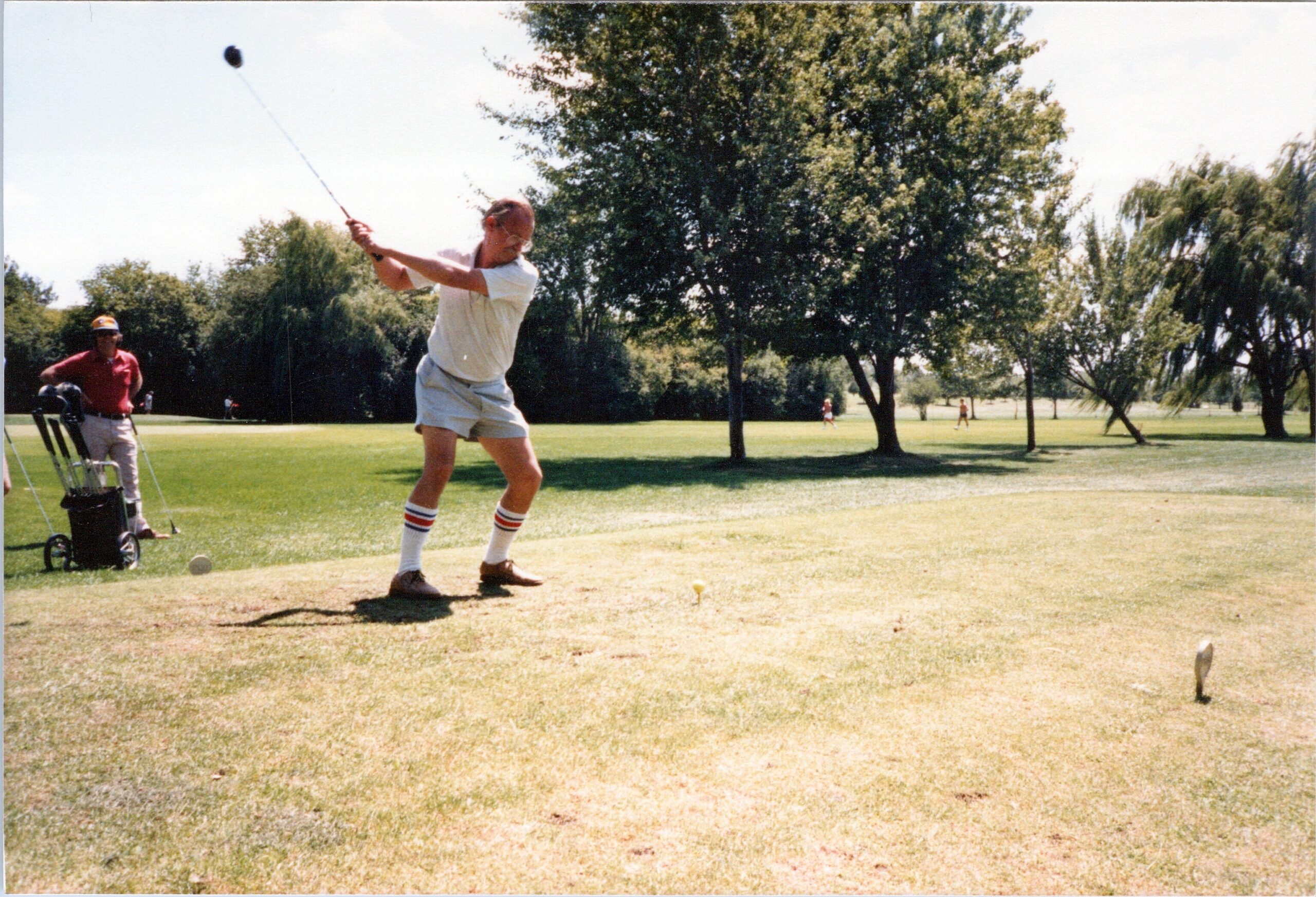 Golfer in white shirt and striped socks swings a club on a sunny course while a caddie with a cart watches nearby.