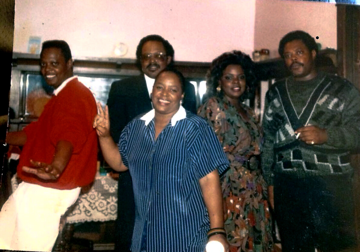 Five adults posing for a group photo indoors; from left to right: man in red shirt, man in suit, woman in blue striped dress, woman in floral dress, man in a patterned sweater.