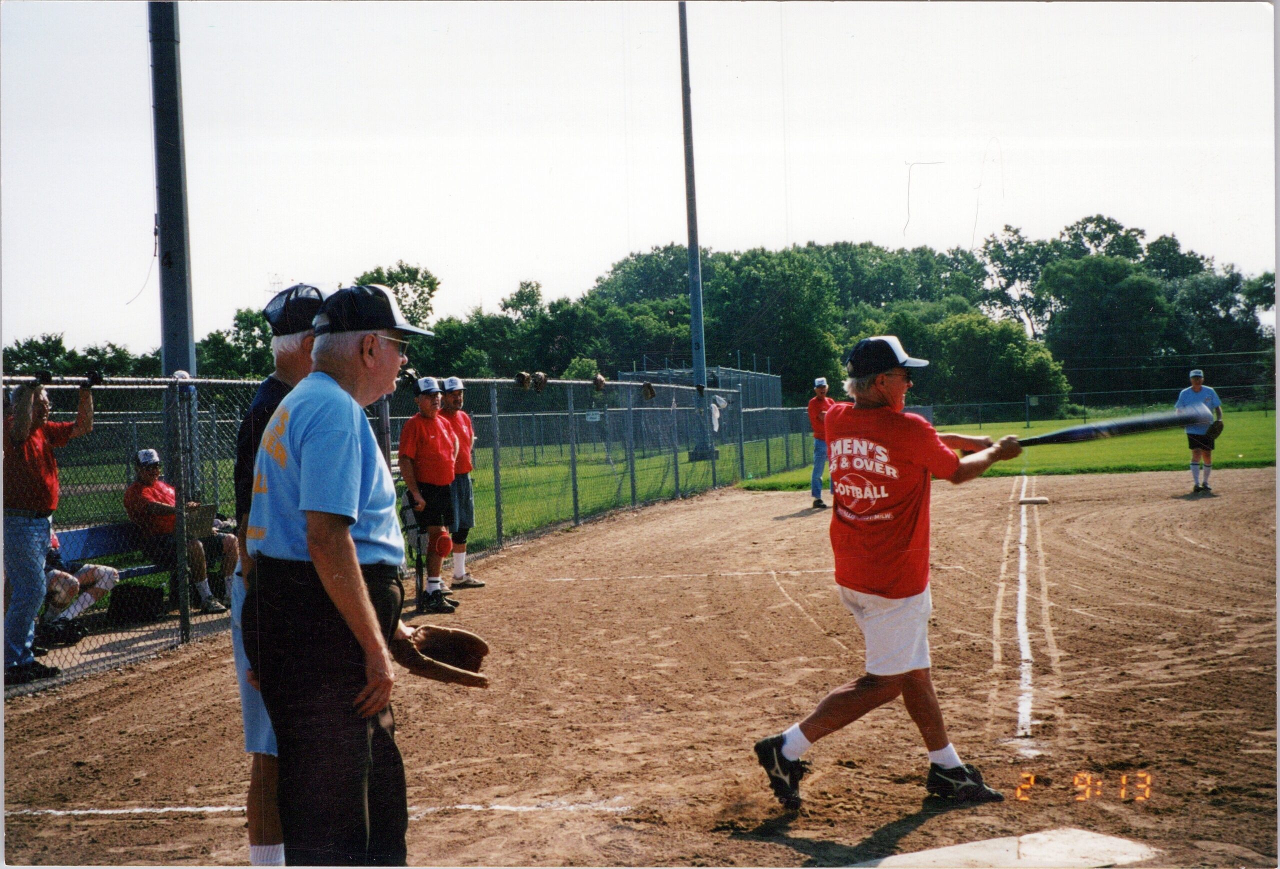 Softball game in progress on a dusty field; a batter in a red shirt swings as spectators watch from behind a chain-link fence.