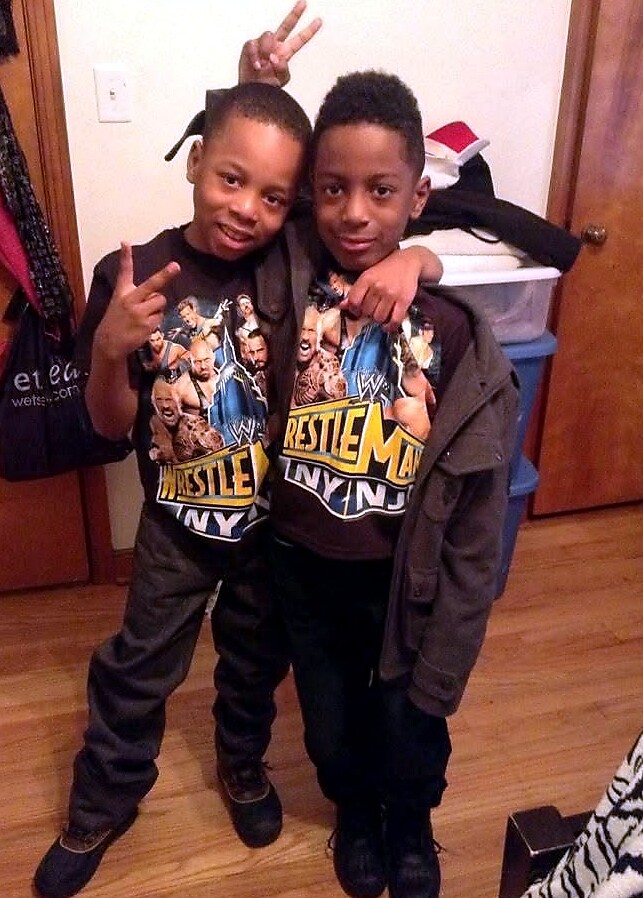 Two young boys pose together indoors; one makes a peace sign while the other has an arm around him, both wearing WrestleMania shirts.