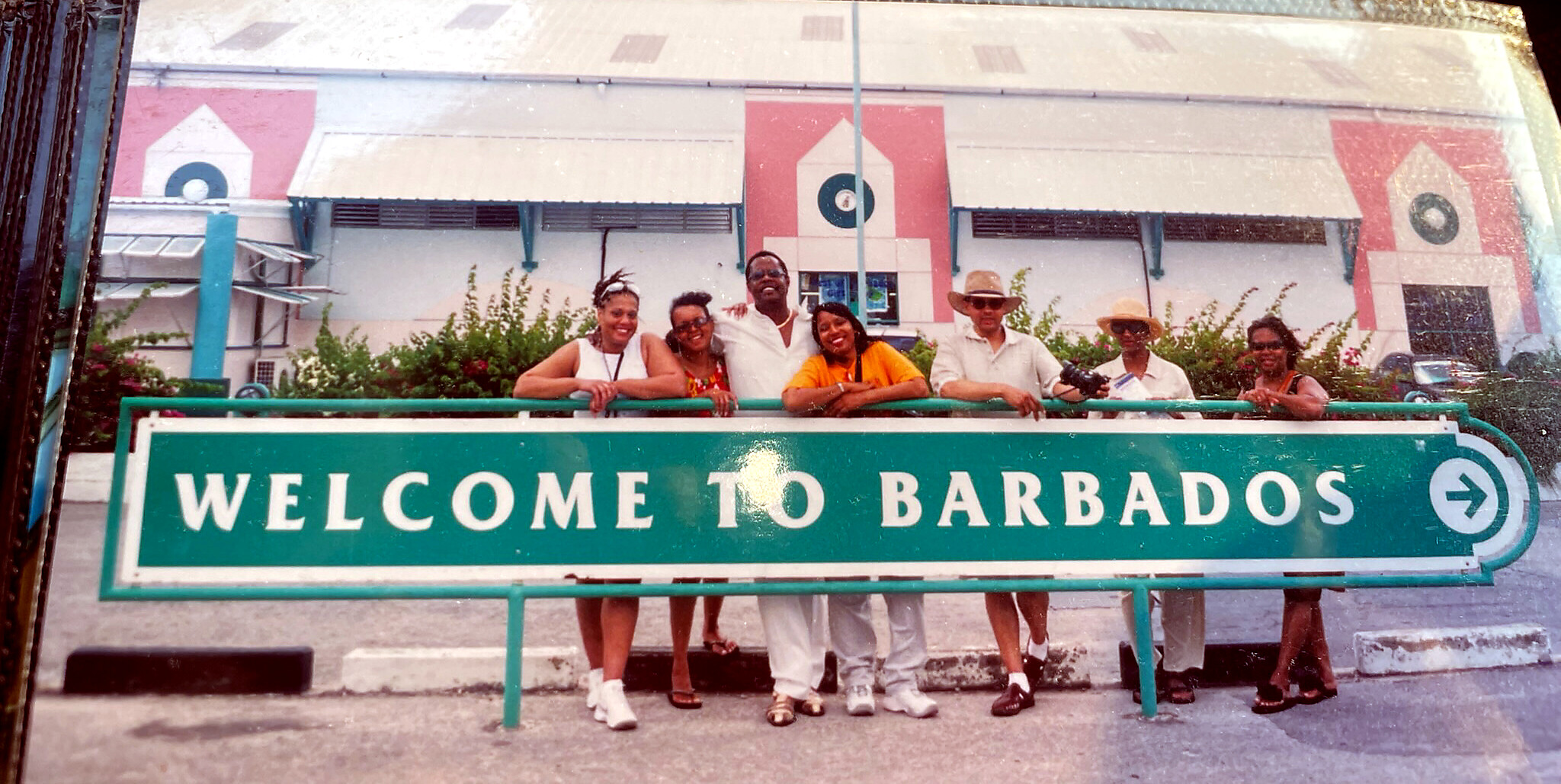 Group of travelers posing behind a green 'Welcome to Barbados' sign at a border/entrance point, smiling for the camera.
