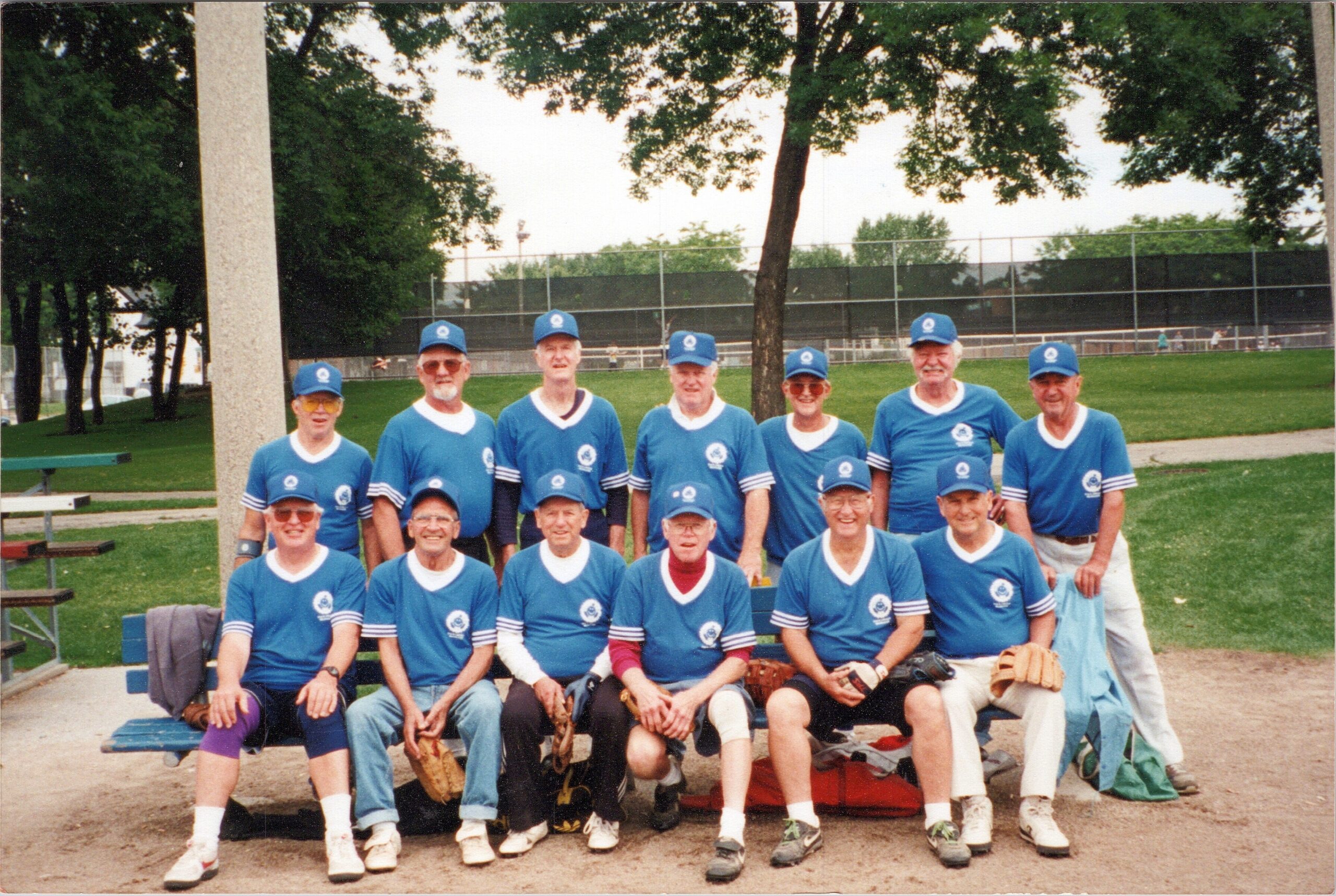 A group of older men in matching blue baseball jerseys and caps posing for a team photo outdoors in a park, with a bench and trees in the background.