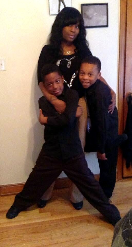 Woman standing behind two boys who hug her legs, all dressed in black in a home hallway setting with framed pictures on the wall nearby.