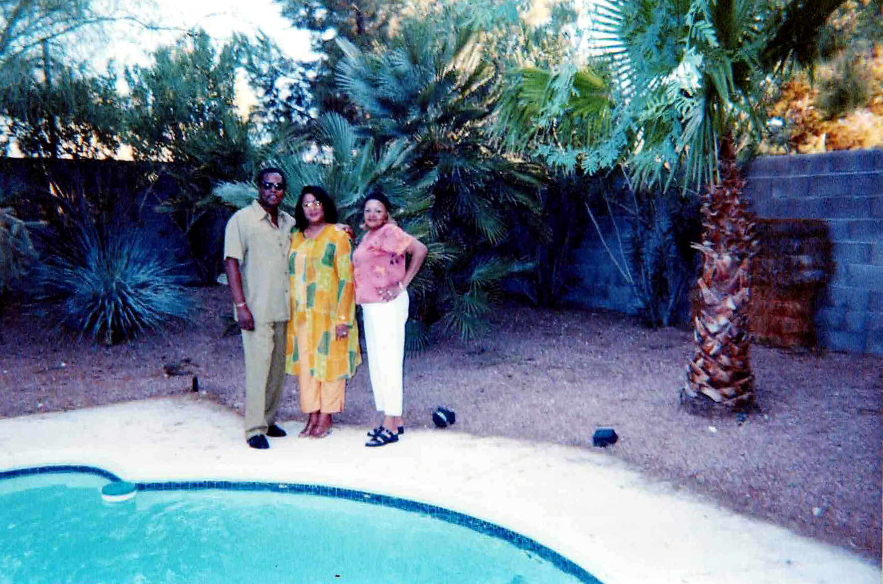 Three adults posing by a pool in a desert backyard, with tall palm plants in the background; man in beige, two women in yellow and pink outfits.