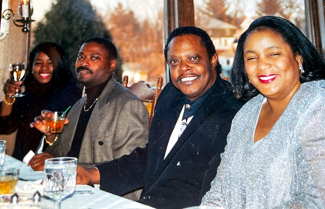 A group of four adults seated at a dining table, toasting with glasses and smiling at the camera in a formal setting.