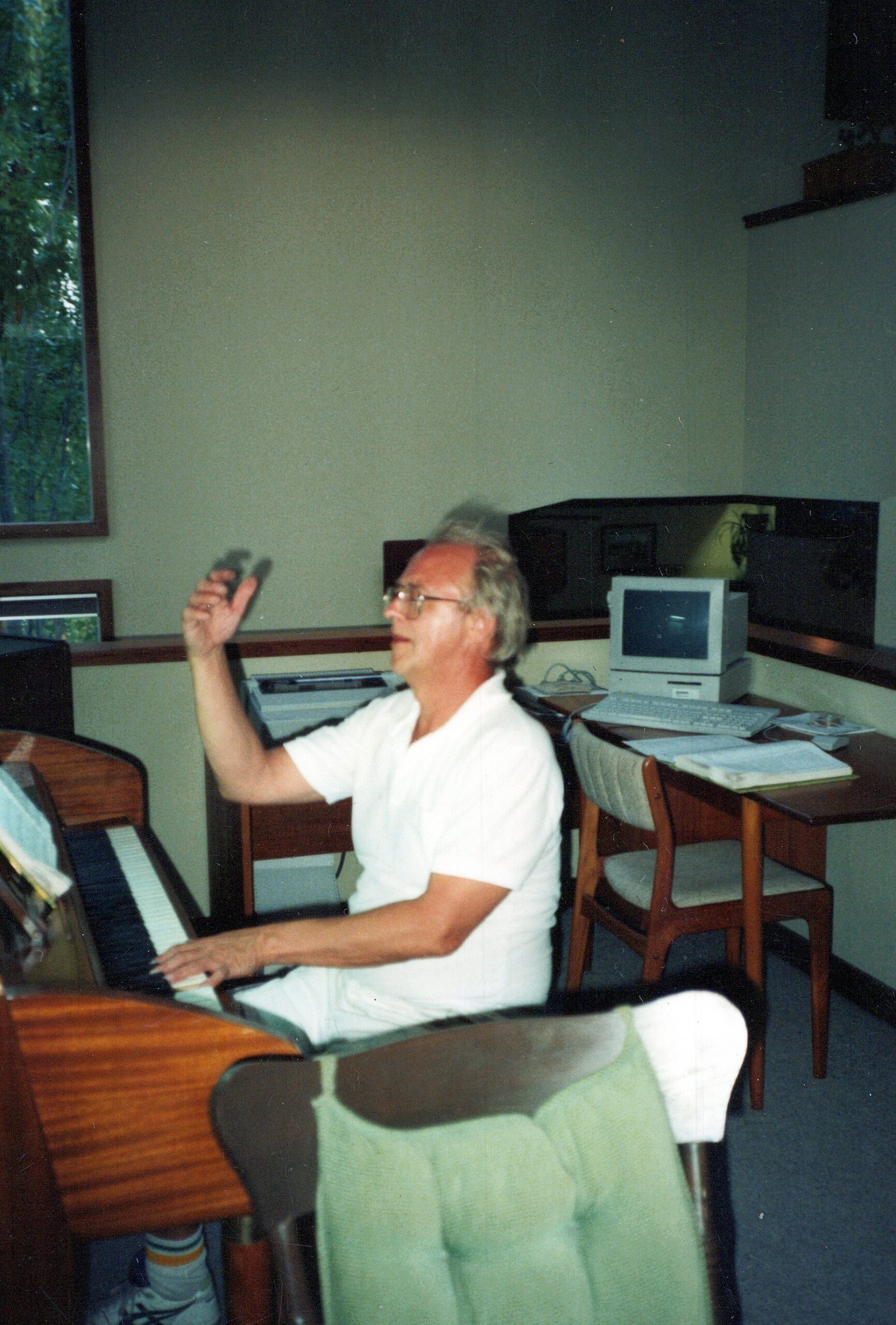 Older man in a white shirt sits at a piano, raising his right hand mid-play in a small office or music room with a computer and papers on a desk behind him.