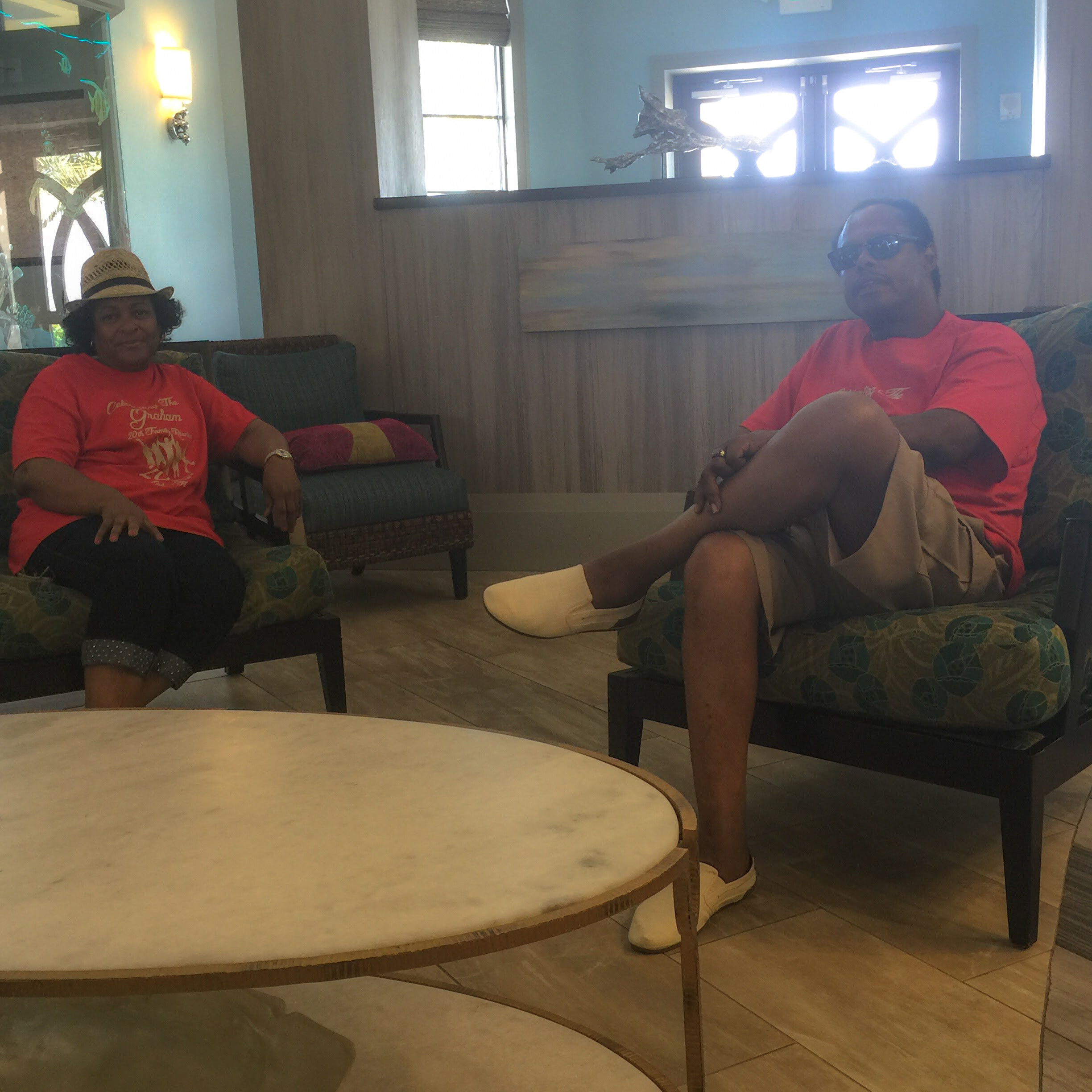 Two people in red shirts sit on couches in a lounge area, round table in foreground, wearing relaxed expressions.