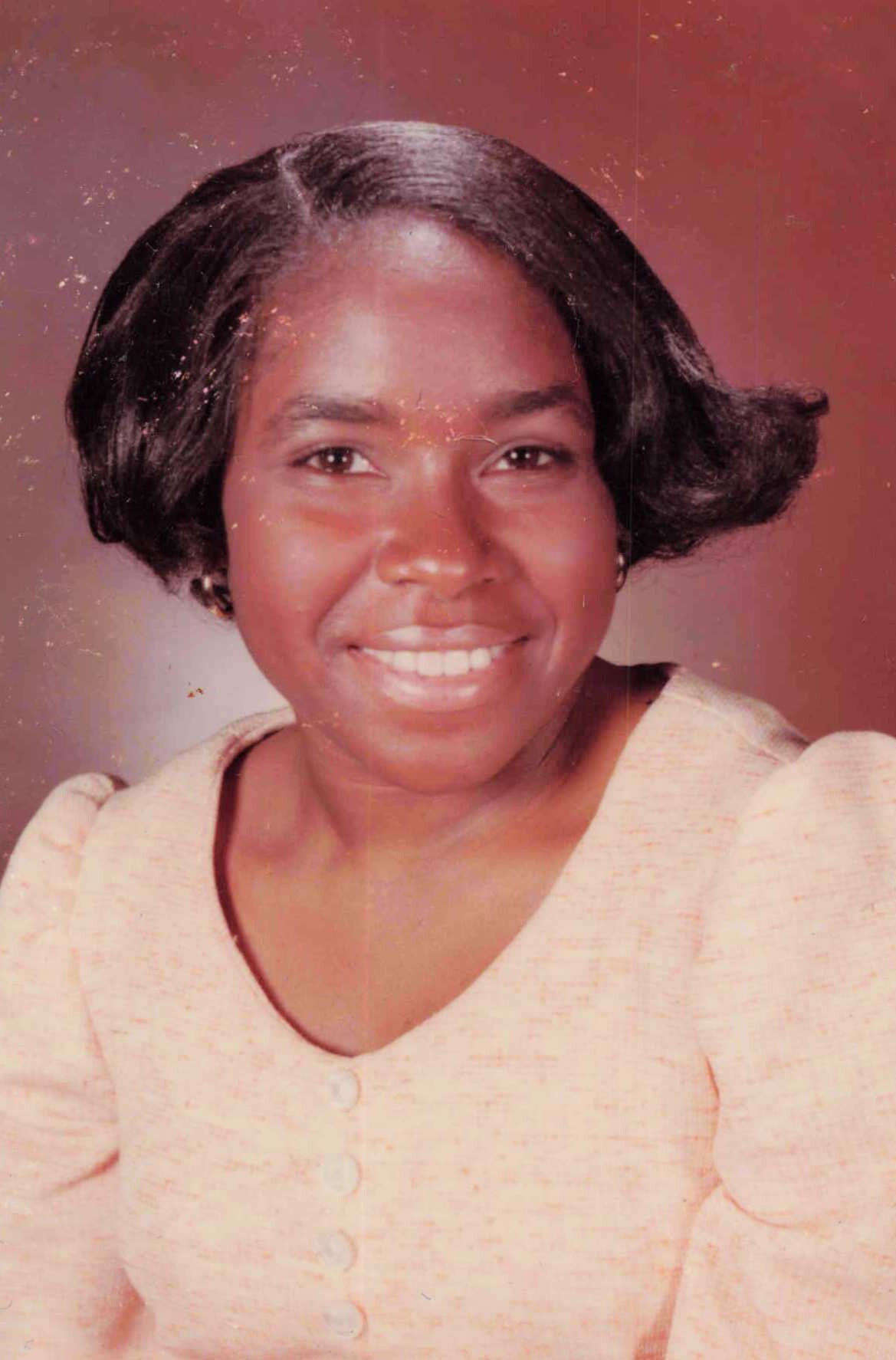 Close-up portrait of a smiling woman with short, dark hair wearing a peach cardigan against a warm backdrop.