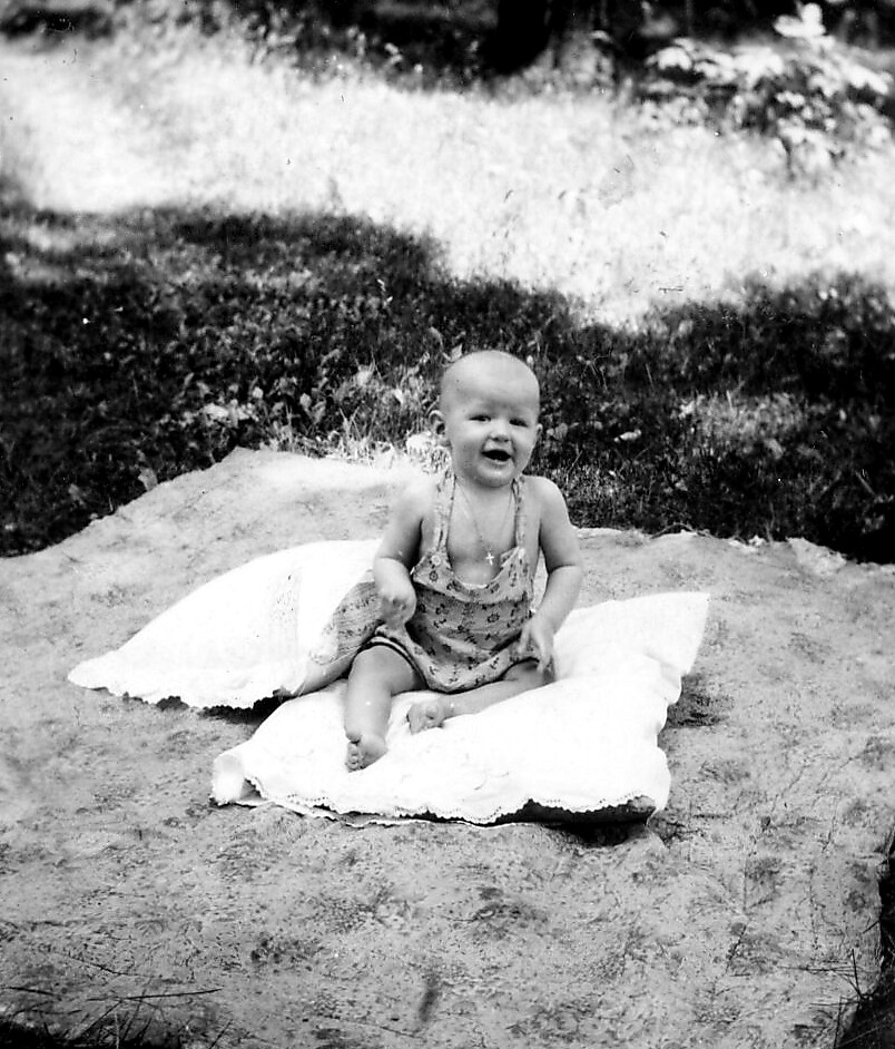 Baby sits on a white towel on a sandy beach, smiling while wearing a one-piece swimsuit and necklace.