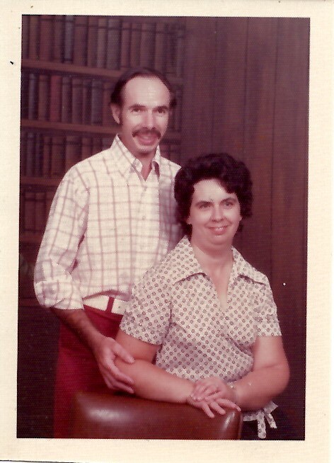 Smiling couple posing for a portrait: a man stands behind a seated woman with his hand on the chair, in a room with a bookshelf backdrop.