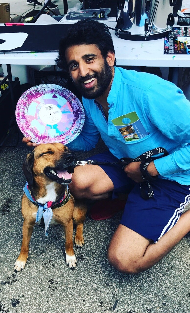 Man in a blue shirt kneels beside a happy brown dog wearing a bandana at an outdoor event, holding a bright tie-dye paper plate.