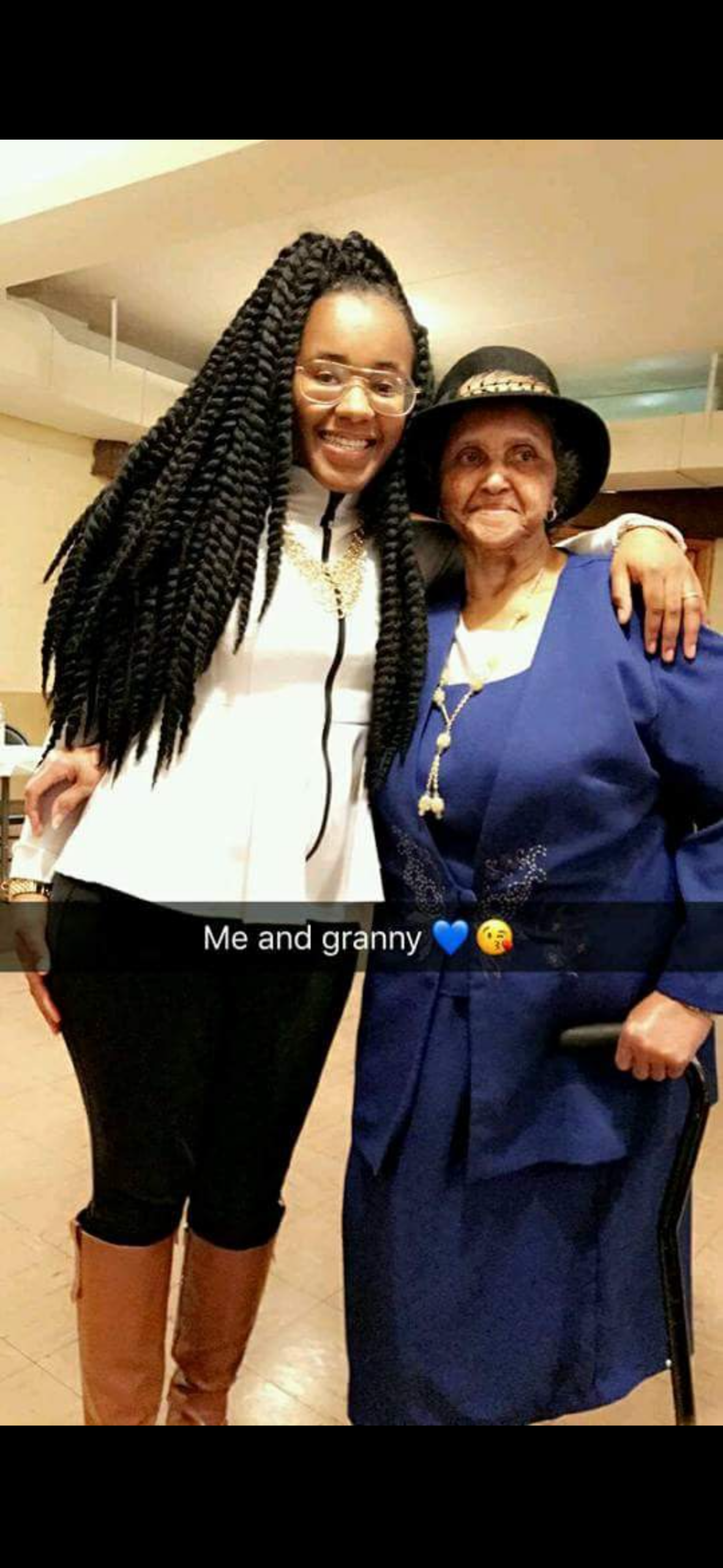 Young woman with long braided hair and glasses posing with her granny in a blue outfit indoors, smiling together.