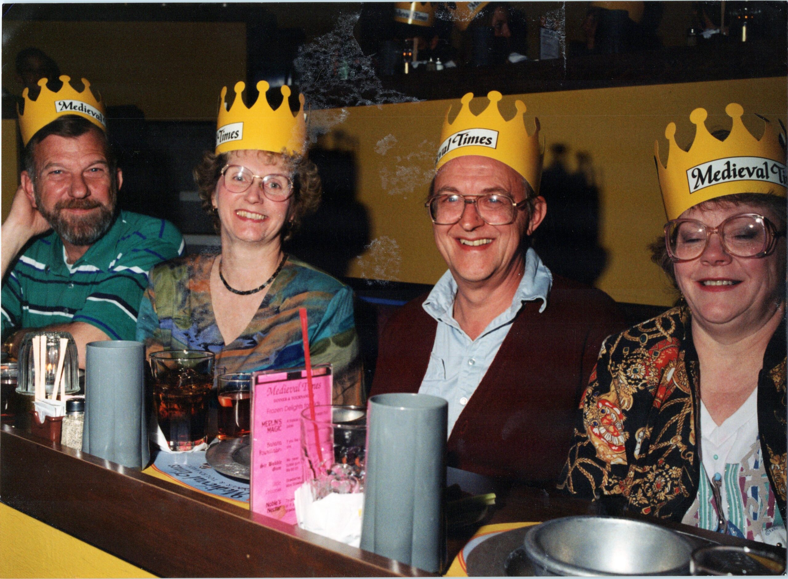Four smiling adults wearing yellow Medieval Times crowns sit at a restaurant table with drinks and menus in front of them.