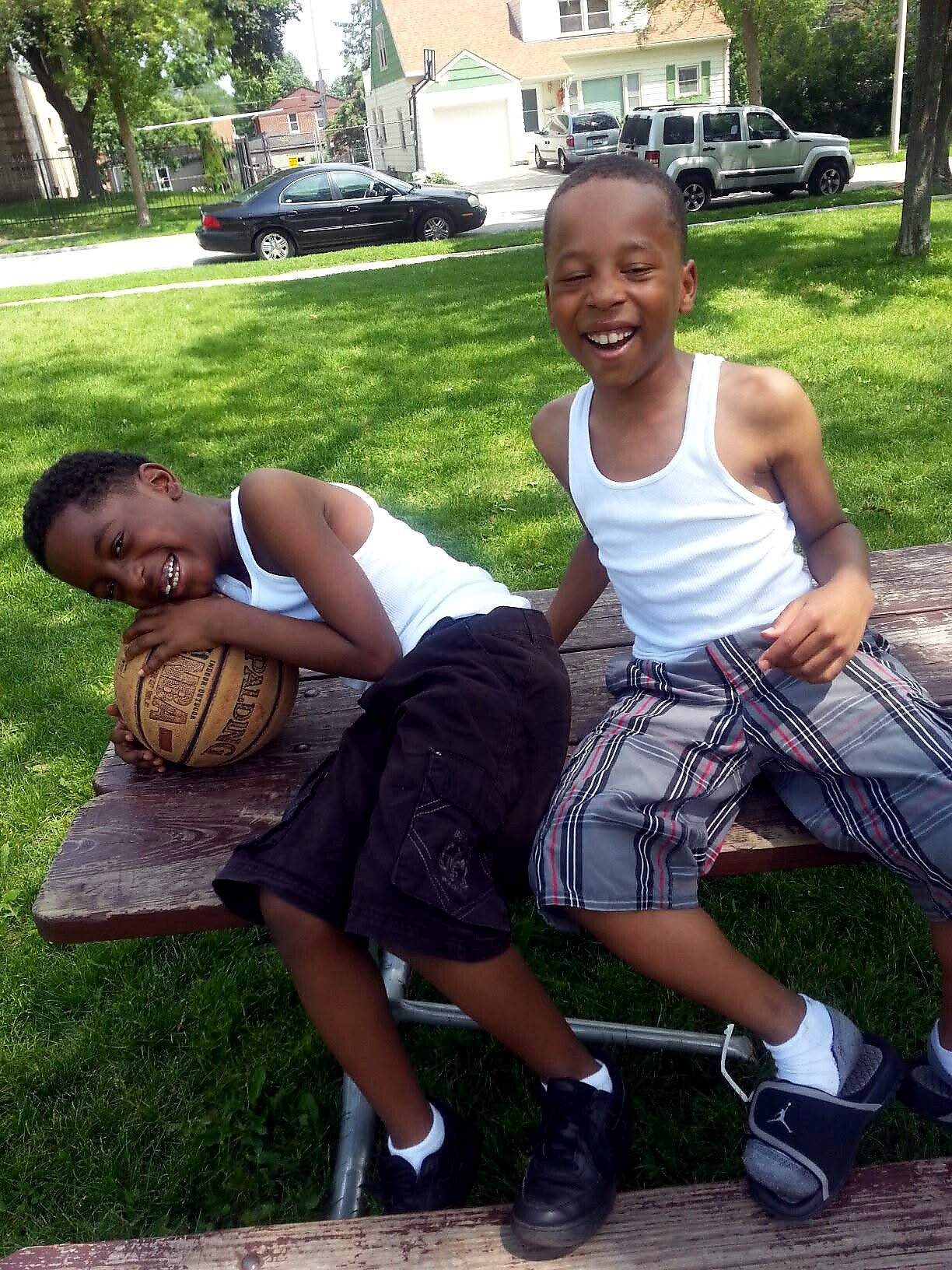 Two boys in white tank tops laughing on a park bench, one resting his head on a basketball while the other leans back and smiles.