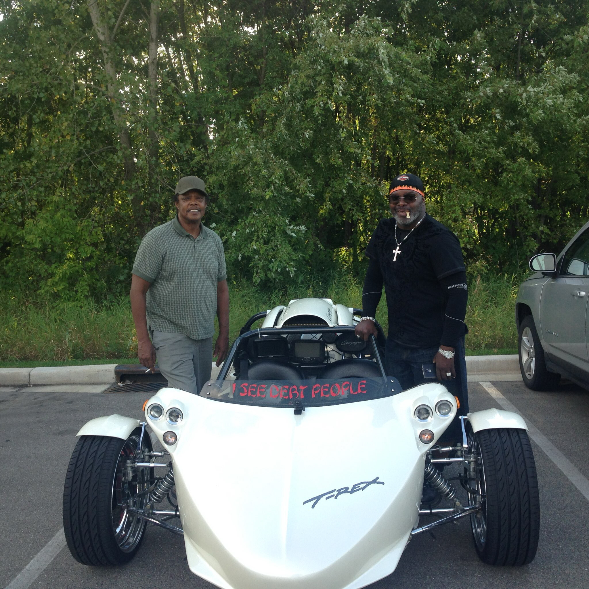 Two men stand beside a white three-wheeled motorcycle in a parking lot, with trees in the background.