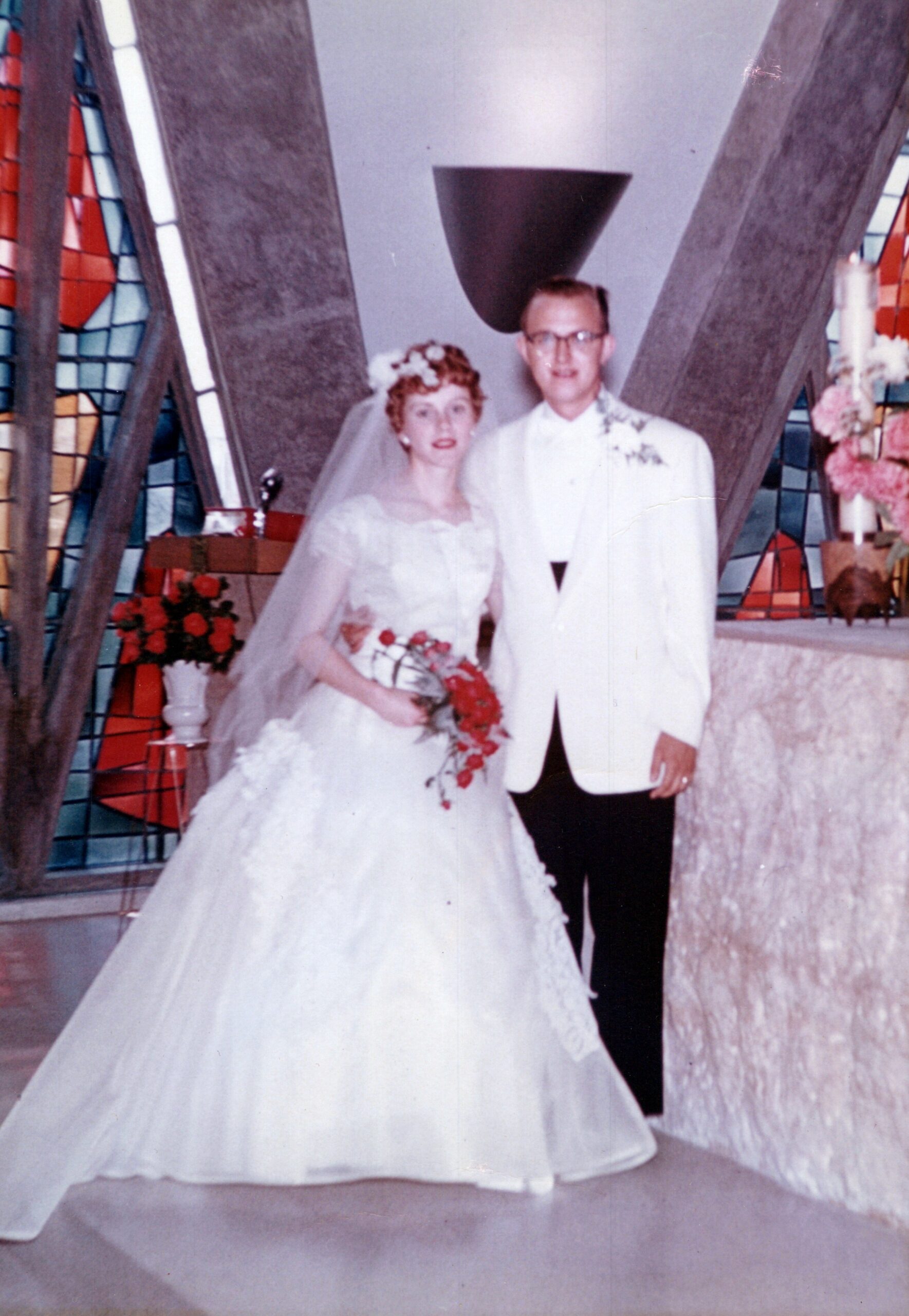 Bride in a white wedding dress holding a bouquet, standing with a groom in a white suit inside a church with colorful stained-glass windows behind them.