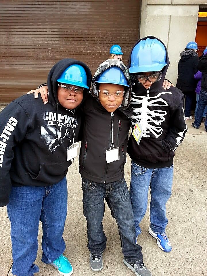 Three children standing with arms around each other, wearing blue safety helmets outside a building.