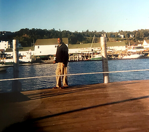 Man standing on a wooden dock by a calm harbor, with boats and white buildings in the background.