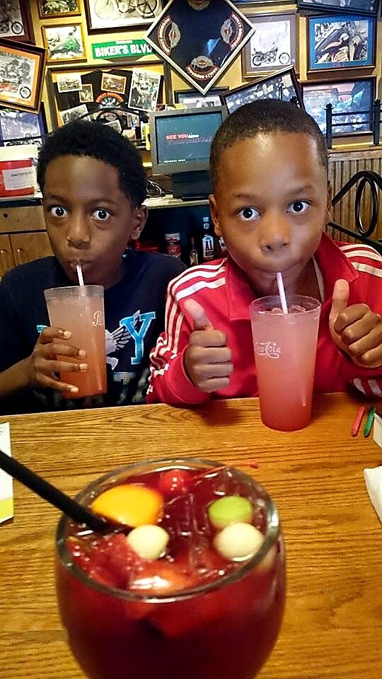 Two boys drink pink beverages through straws at a restaurant, with a big fruit punch in the foreground.