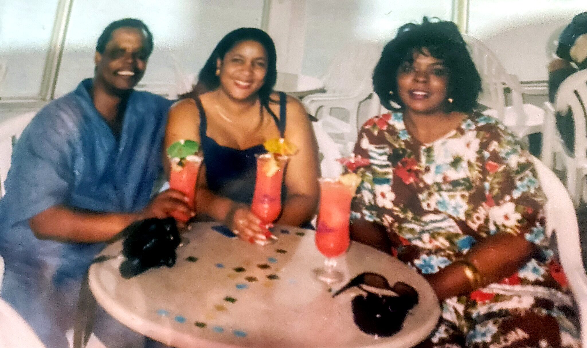Three friends sitting at a table, smiling, each with a bright tropical drink garnished with fruit.
