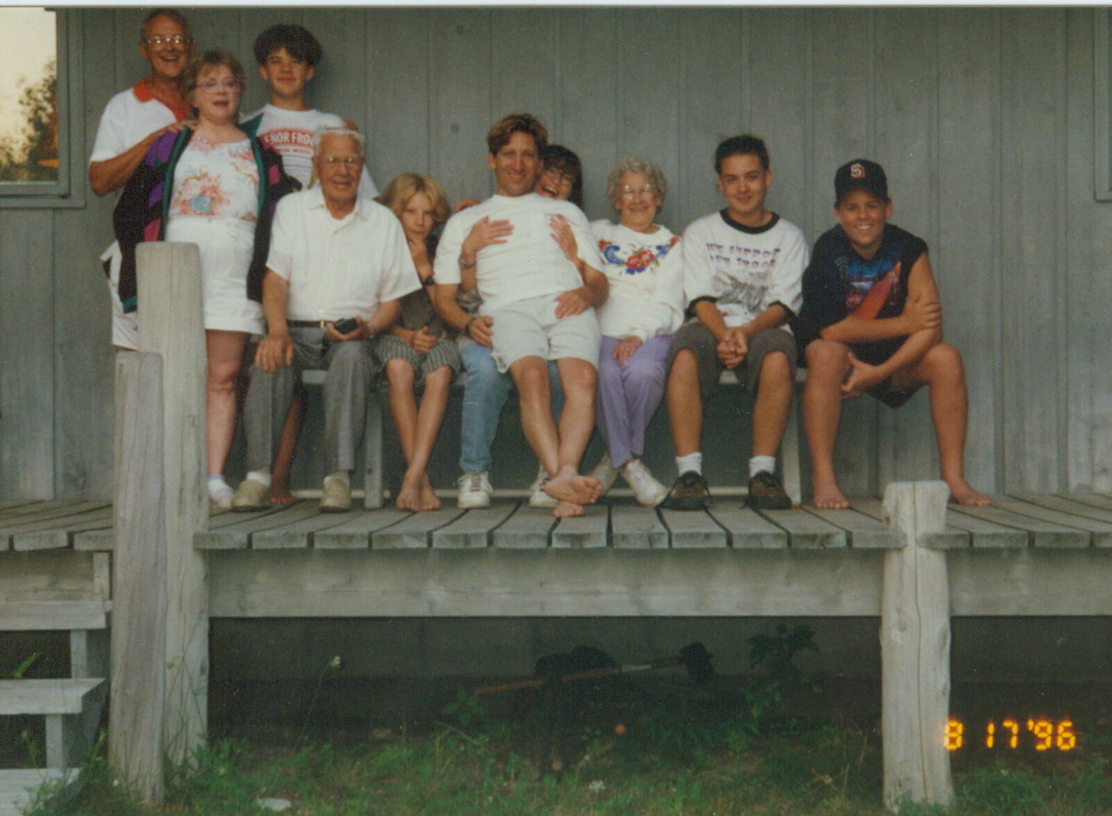 Group of twelve people posing together on a weathered wooden deck in front of a gray wall, smiling for a family photo.