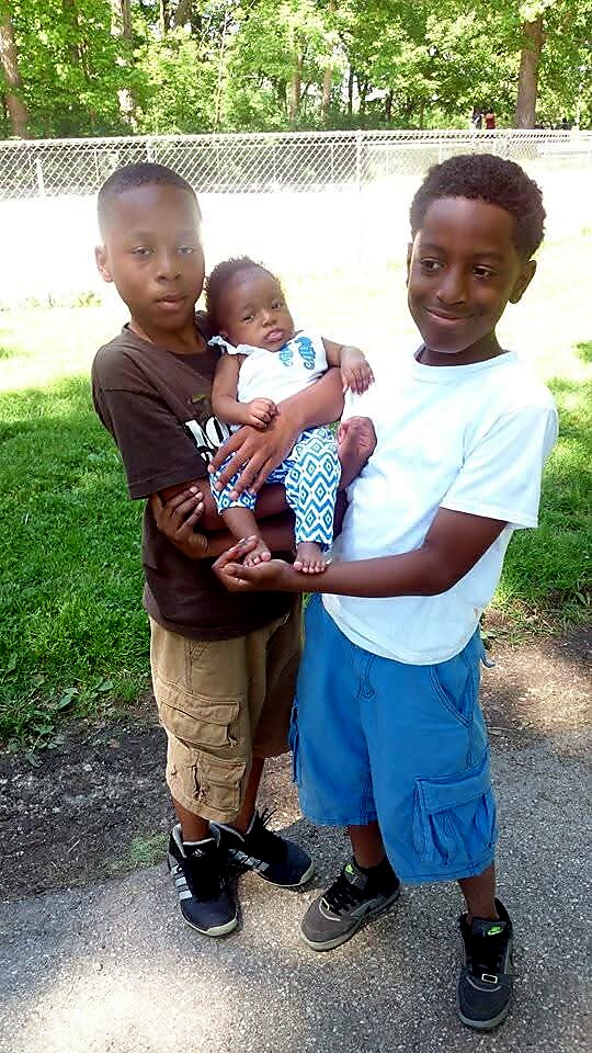 Three kids outdoors, two older brothers gently hold a baby between them in a grassy area with a chain-link fence behind.