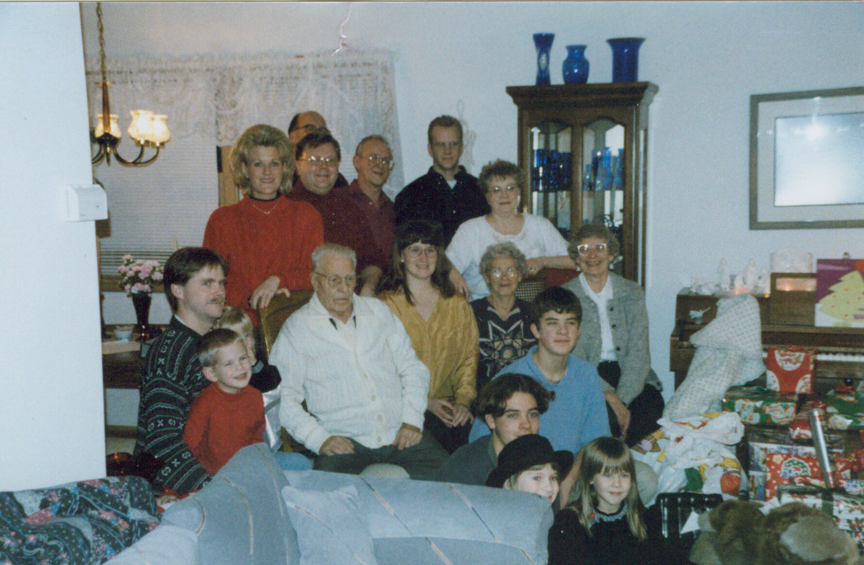 Multi-generation family group photo in a living room during a Christmas celebration, with wrapped gifts in the foreground and everyone smiling at the camera.