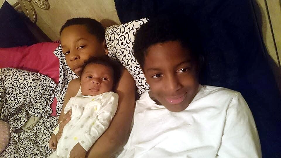 Three siblings cuddling on a bed: a baby held by the middle child, with an older boy on the left and a teen smiling on the right, surrounded by patterned pillows.