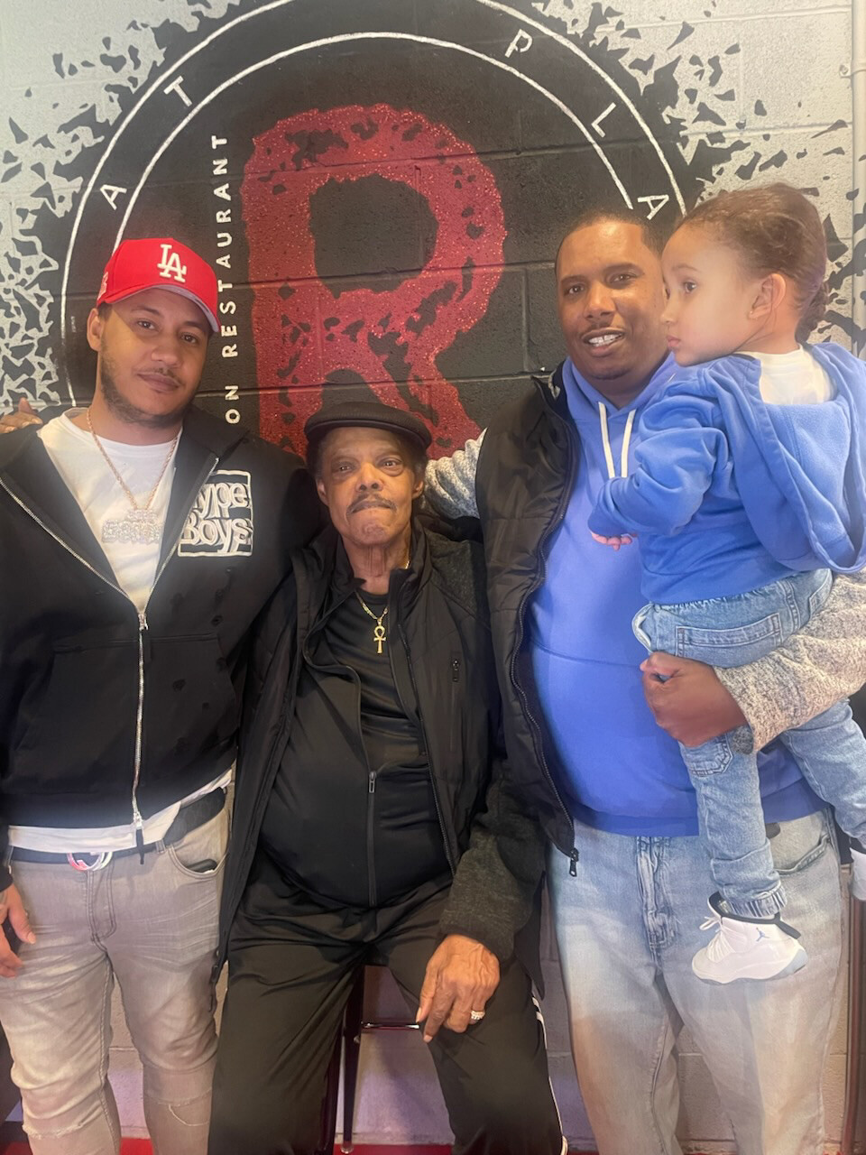 Group photo in a restaurant: an elderly man in black with a hat sits center, flanked by three people—a man in a red LA cap on the left and a man in a blue hoodie holding a child on the right.