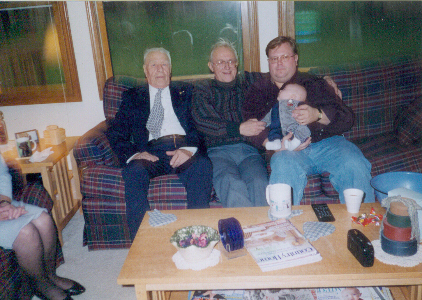 Three generations on a plaid sofa: an elderly man in a suit, a smiling senior, and a younger man holding a baby with a cozy living room backdrop.