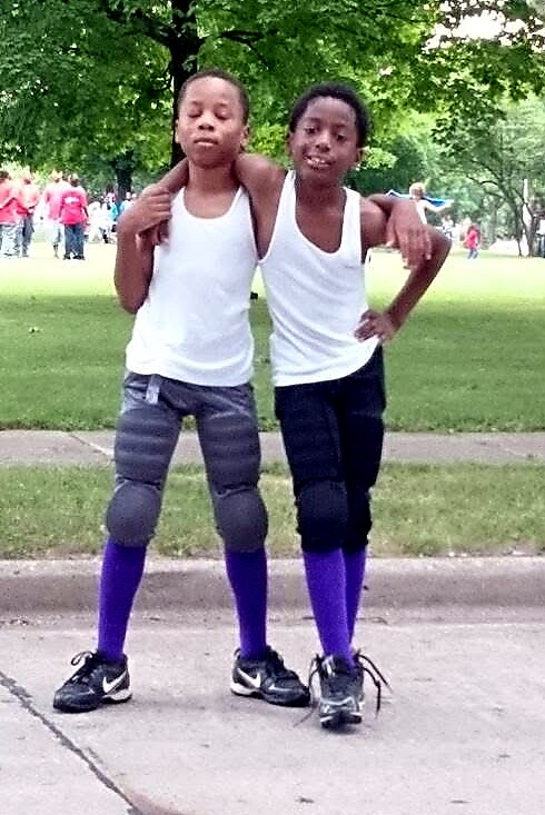 Two boys in white tank tops pose with arms around each other in a park, wearing purple socks and athletic shoes.