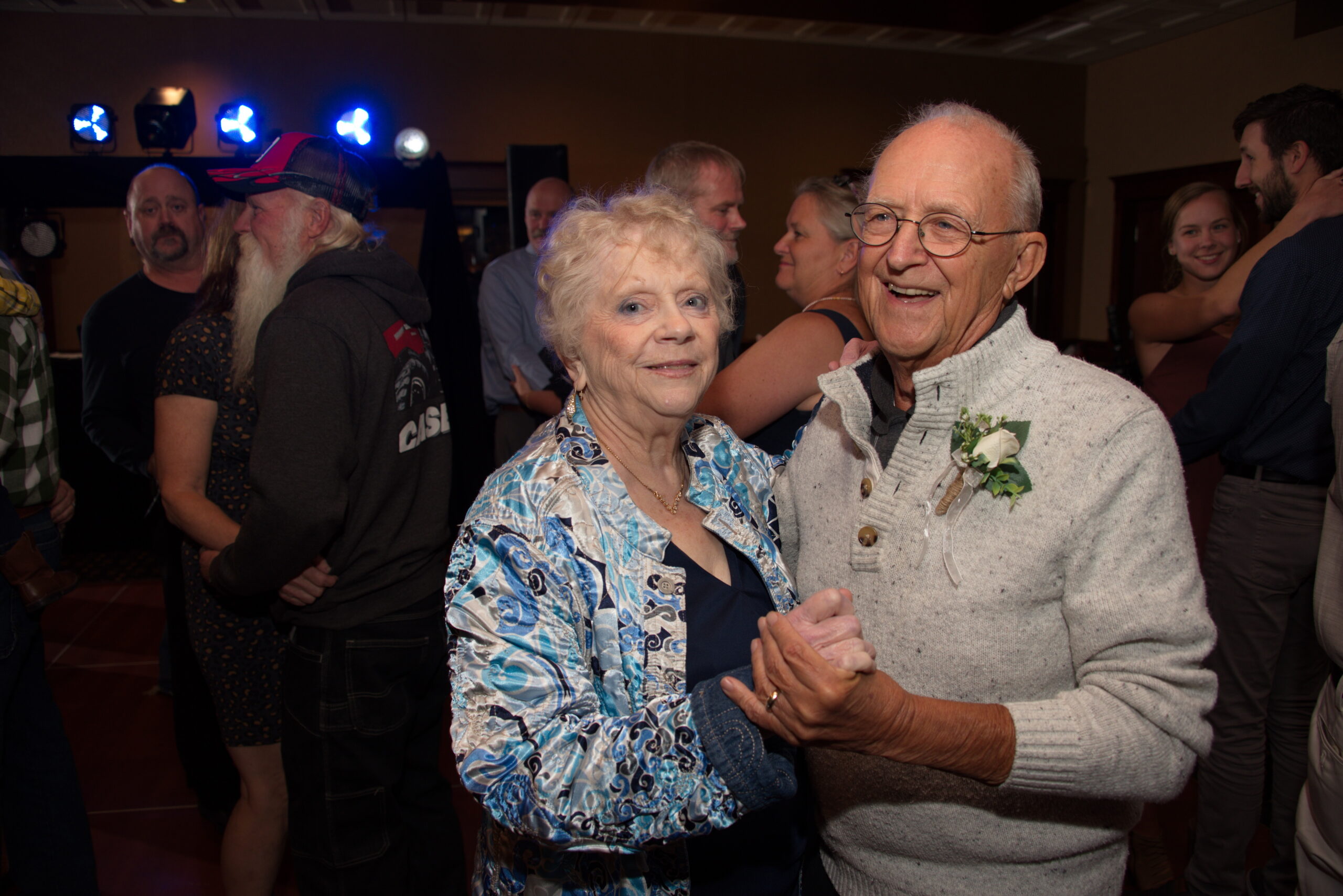 An elderly couple dances together at a social gathering, smiling at the camera with others mingling in the background.