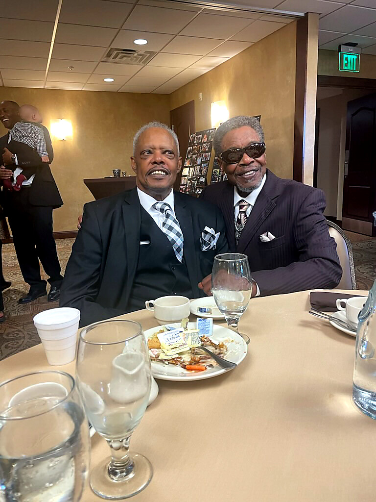 Two men in formal suits pose and smile at a round banquet table with plates, cups, and glasses in a dining hall.