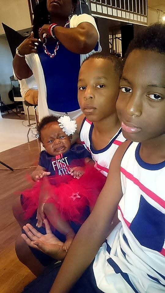 Three children pose for a photo indoors: two older brothers in matching white, blue, and red athletic uniforms and a baby girl in a pink tutu seated on someone's lap in the background.