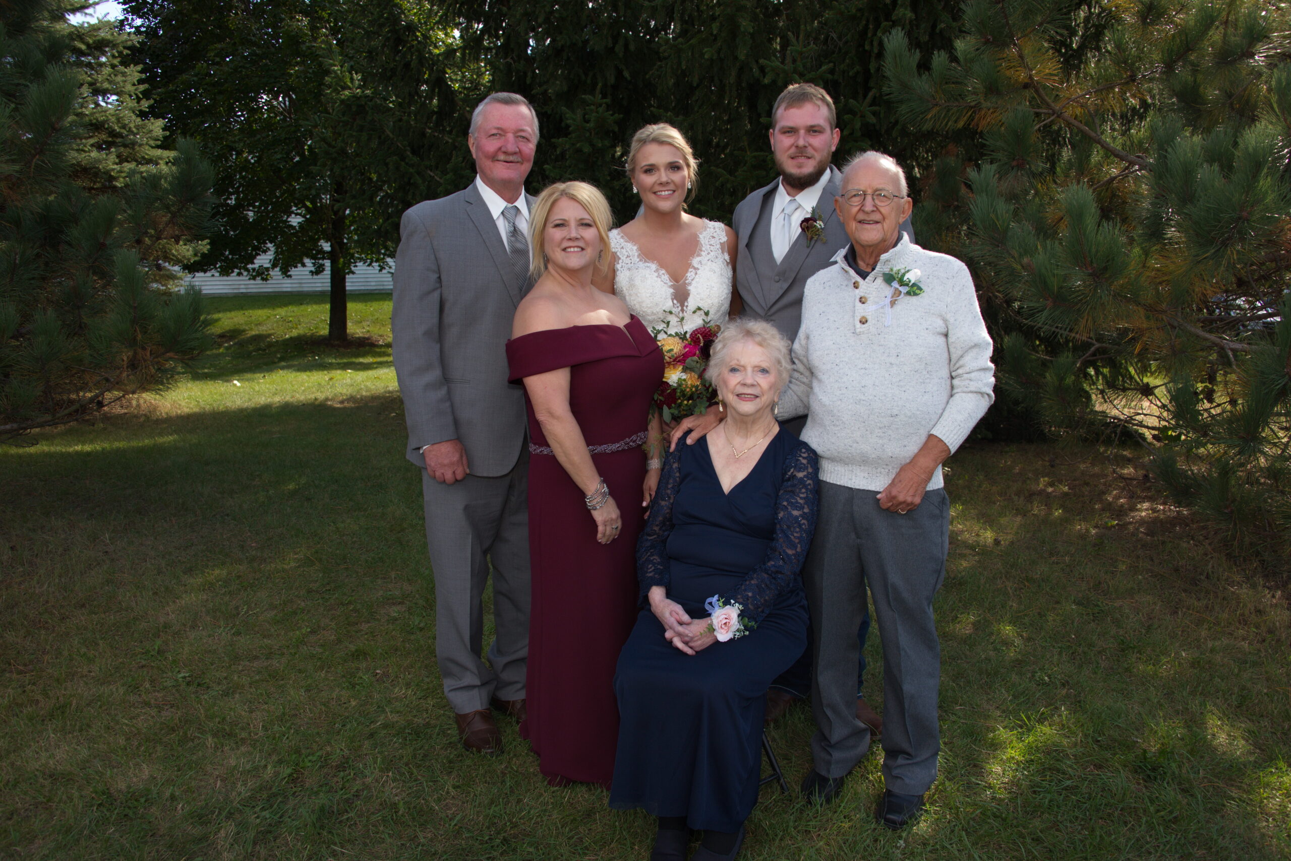 Six family members pose outdoors with trees in the background: bride in a lace white gown, groom in a gray suit, two adults in formal wear, and two elderly relatives (one seated in a navy dress). They smile for the camera on a sunny day at the yard.