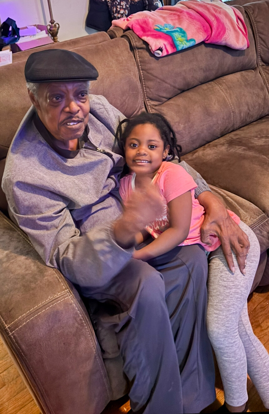 Older man in a cap sits on a brown sofa with a smiling young girl on his lap, they hug warmly.