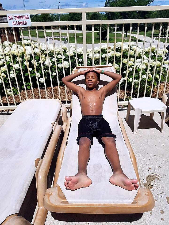 Shirtless boy relaxing on a poolside lounge chair with arms raised over his head; 'No Smoking Allowed' sign and railing behind him.