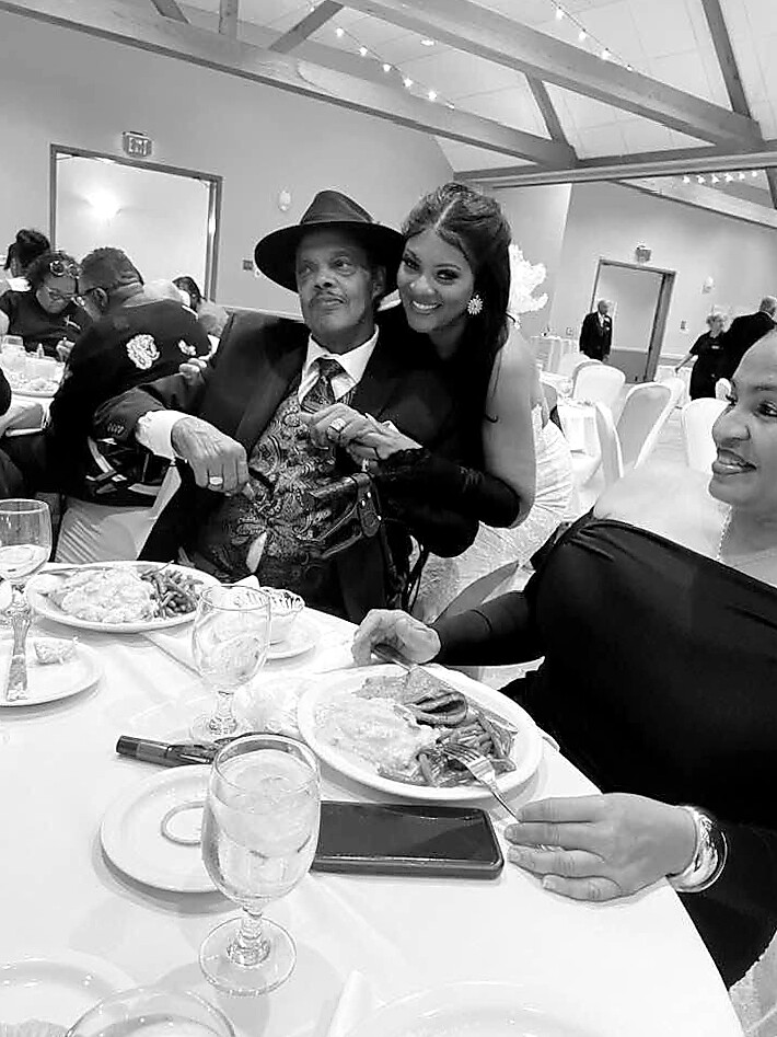 Man in a suit and fedora sitting at a banquet table, smiling as a woman stands behind him with her arm around his shoulder.