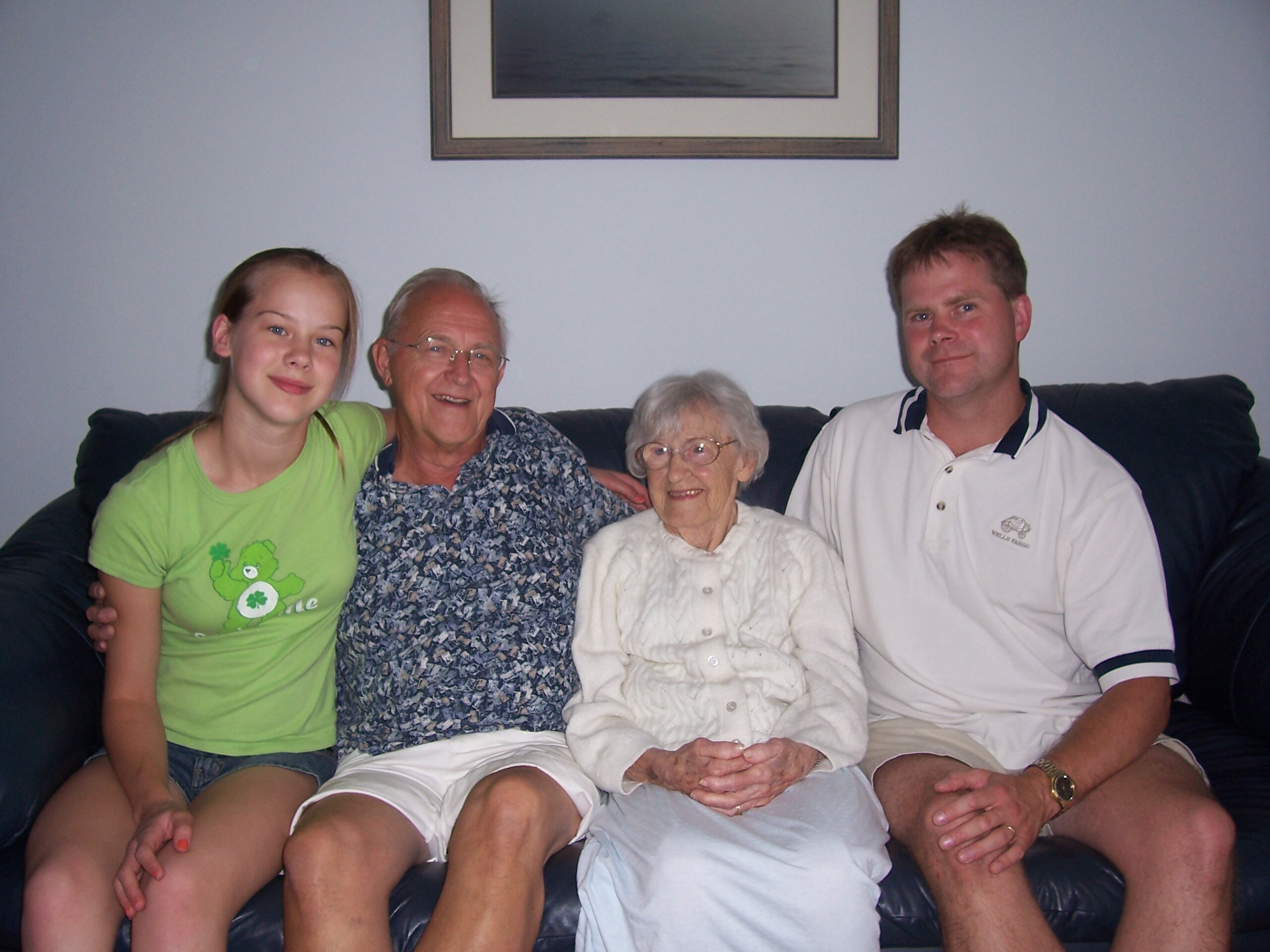 Five people sitting closely on a dark couch in a living room, smiling at the camera against a light blue wall with a framed picture above them.