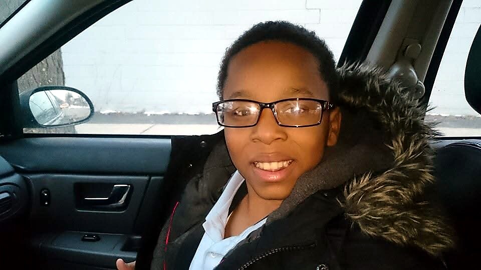Young person wearing glasses sits in a car, smiling at the camera, with a fur-lined hooded coat.