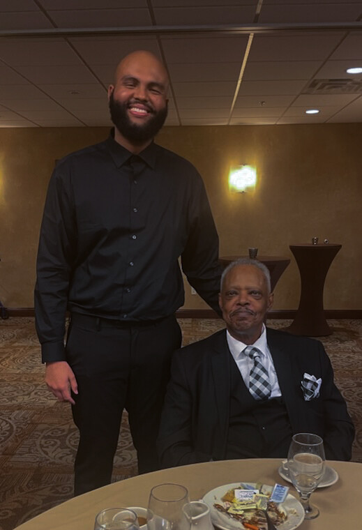 Smiling man in a black shirt stands beside an older gentleman in a suit at a formal event, with a plate and glasses on the table in front.