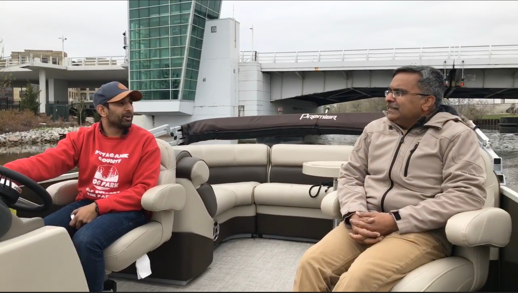 Two men sitting and talking on a beige pontoon boat, with a modern bridge and water in the background.
