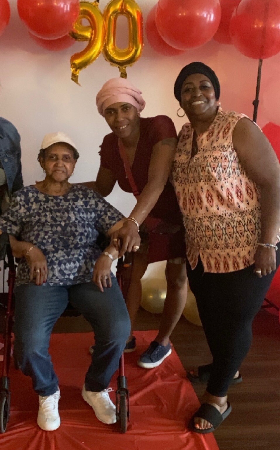 Three women pose together at a 90th birthday party: an elderly woman in a wheelchair flanked by two friends under red and gold balloons, smiling at the camera.