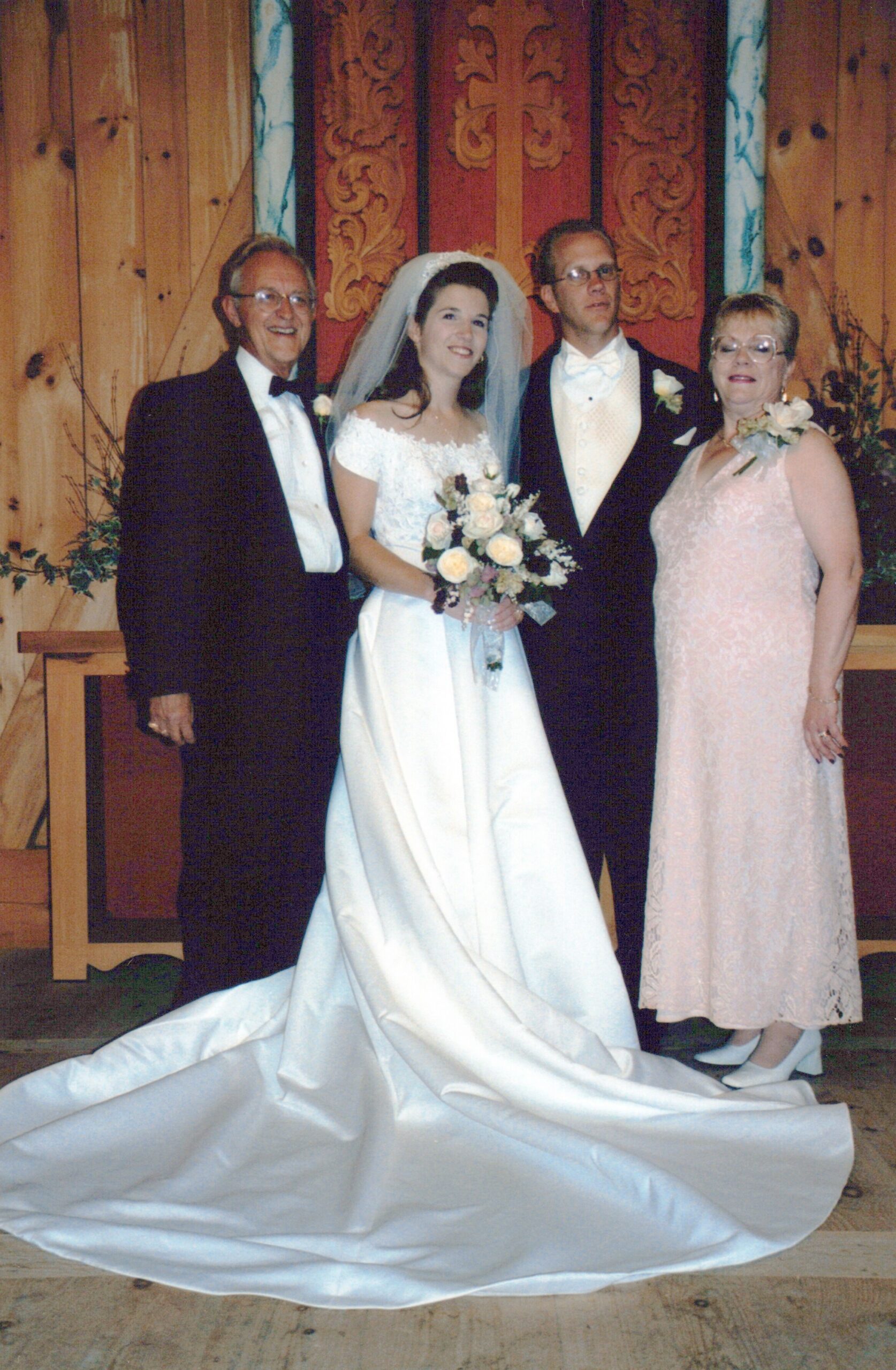 Bride in white gown and veil posing with her parents in formal attire at a wedding ceremony backdrop of carved wooden doors; bouquet in hand.