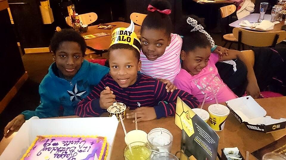Four kids sit at a restaurant table celebrating a birthday, with a frosted cake, colorful drinks, and a birthday crown worn by the center child.