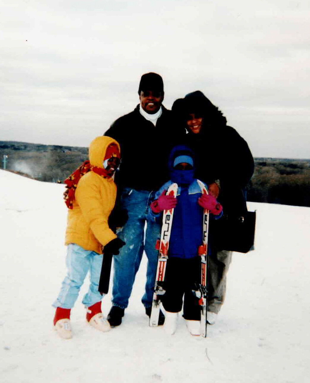 Family of four posing in the snow with ski gear, outdoors on a snowy hill.