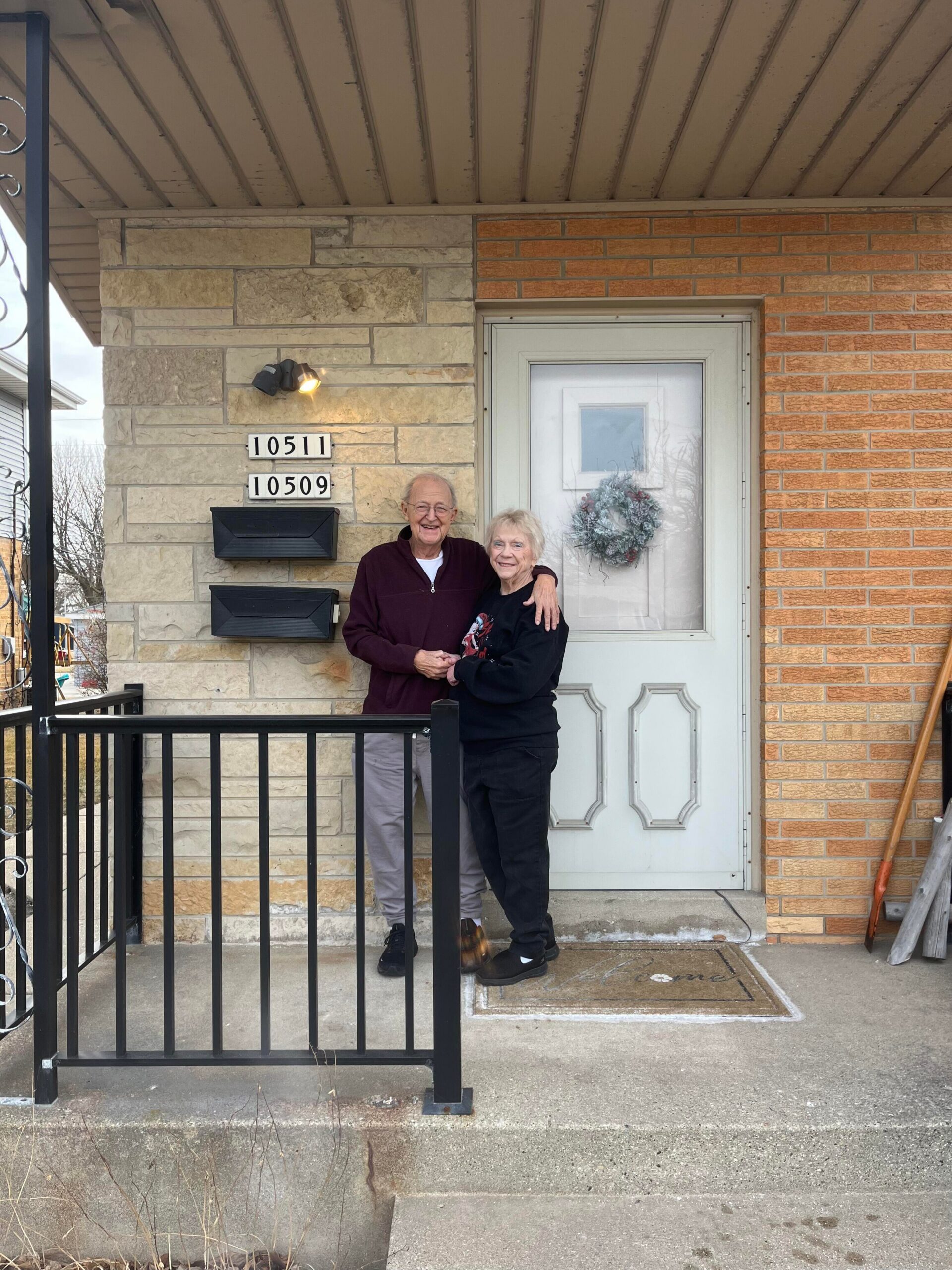 Elderly couple standing arm in arm on a porch in front of a white door with a wreath, stone and brick exterior. Scroll plaques show 10511 and 10509.