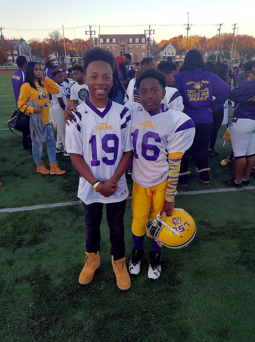 Two young football players in LSU white jerseys (numbers 19 and 16) stand arm in arm on a football field, one holding a yellow LSU helmet; crowd and fall trees in the background.