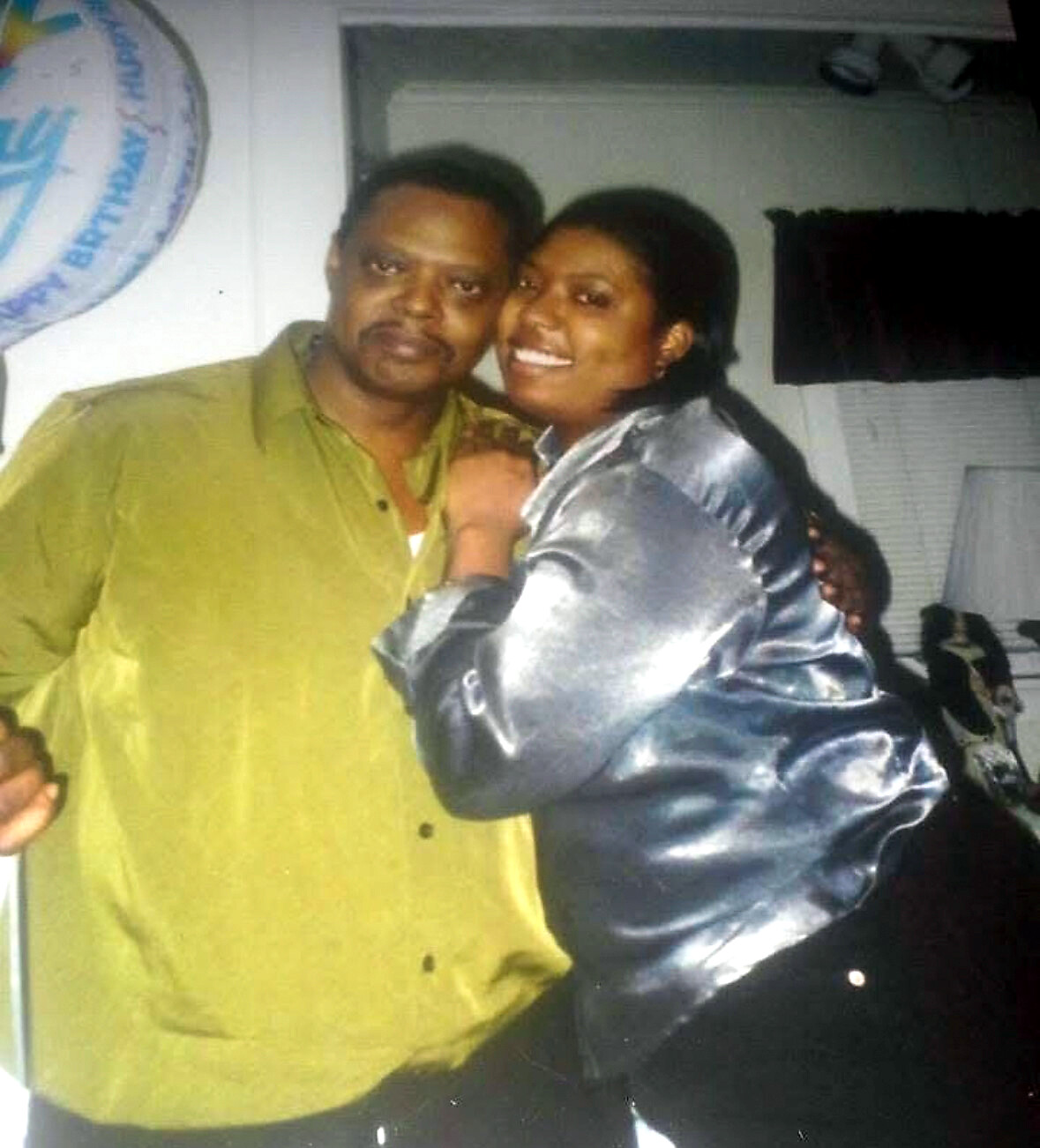 Two people pose closely for a photo: a man in a green shirt and a smiling woman in a shiny jacket, indoors.